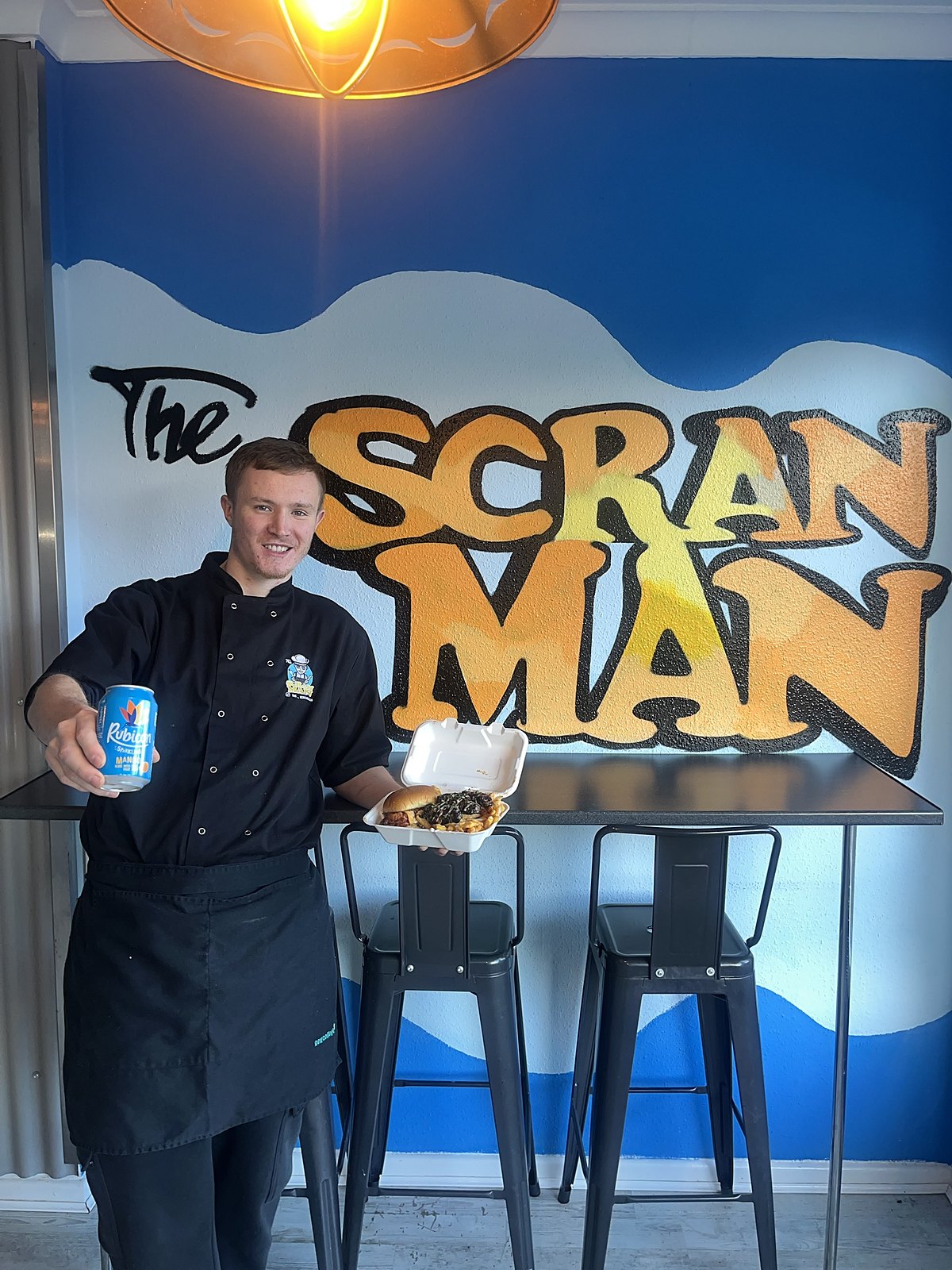 Blake in chef whites holding a meal and a drink in front of The Scran Man mural